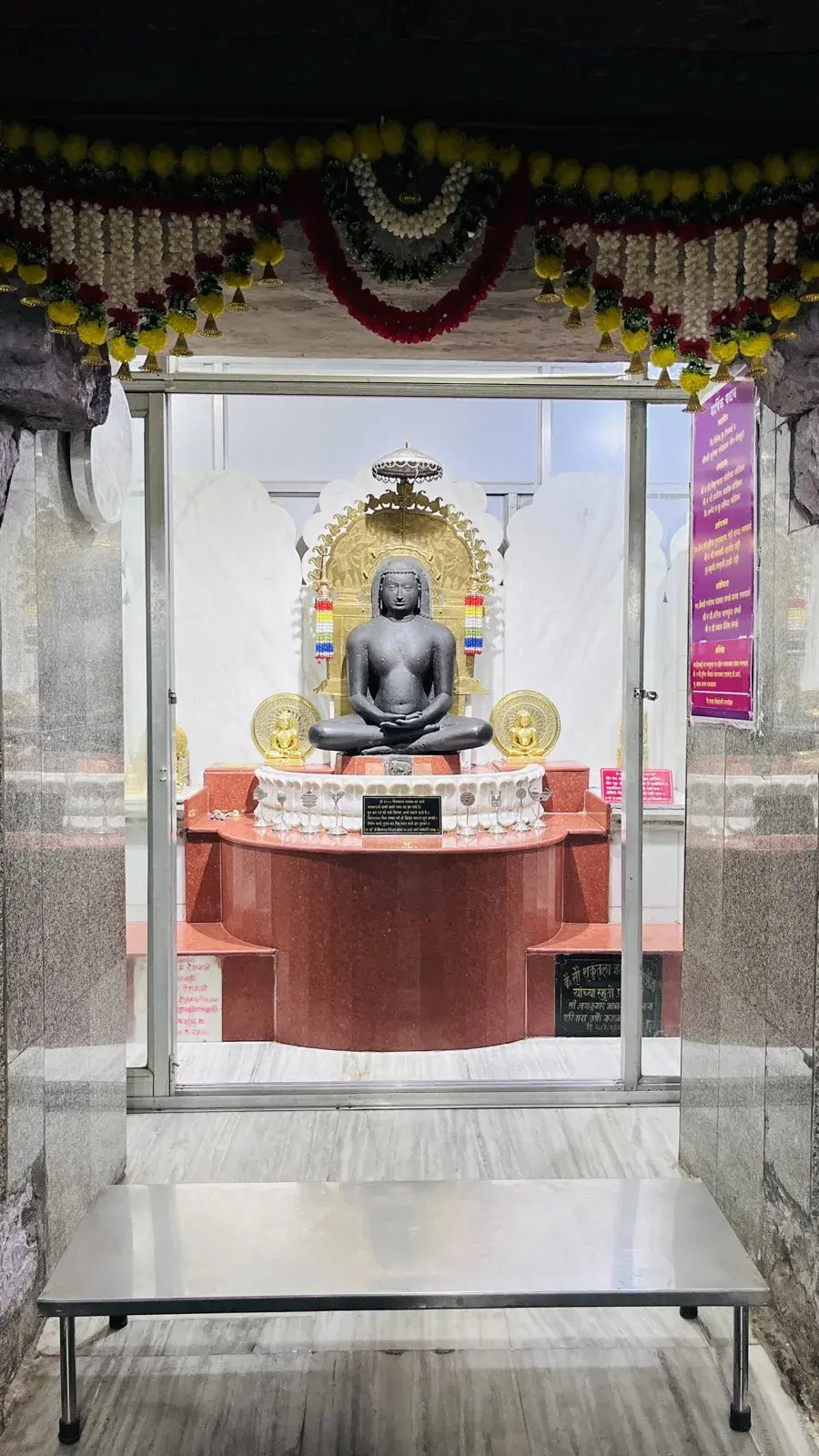 The Moolnayak Bhagwan Vimalnath, a black stone murti seated in Padmasana in the garbhagriha, garlanded and set against a gold prabhavali and chatra.