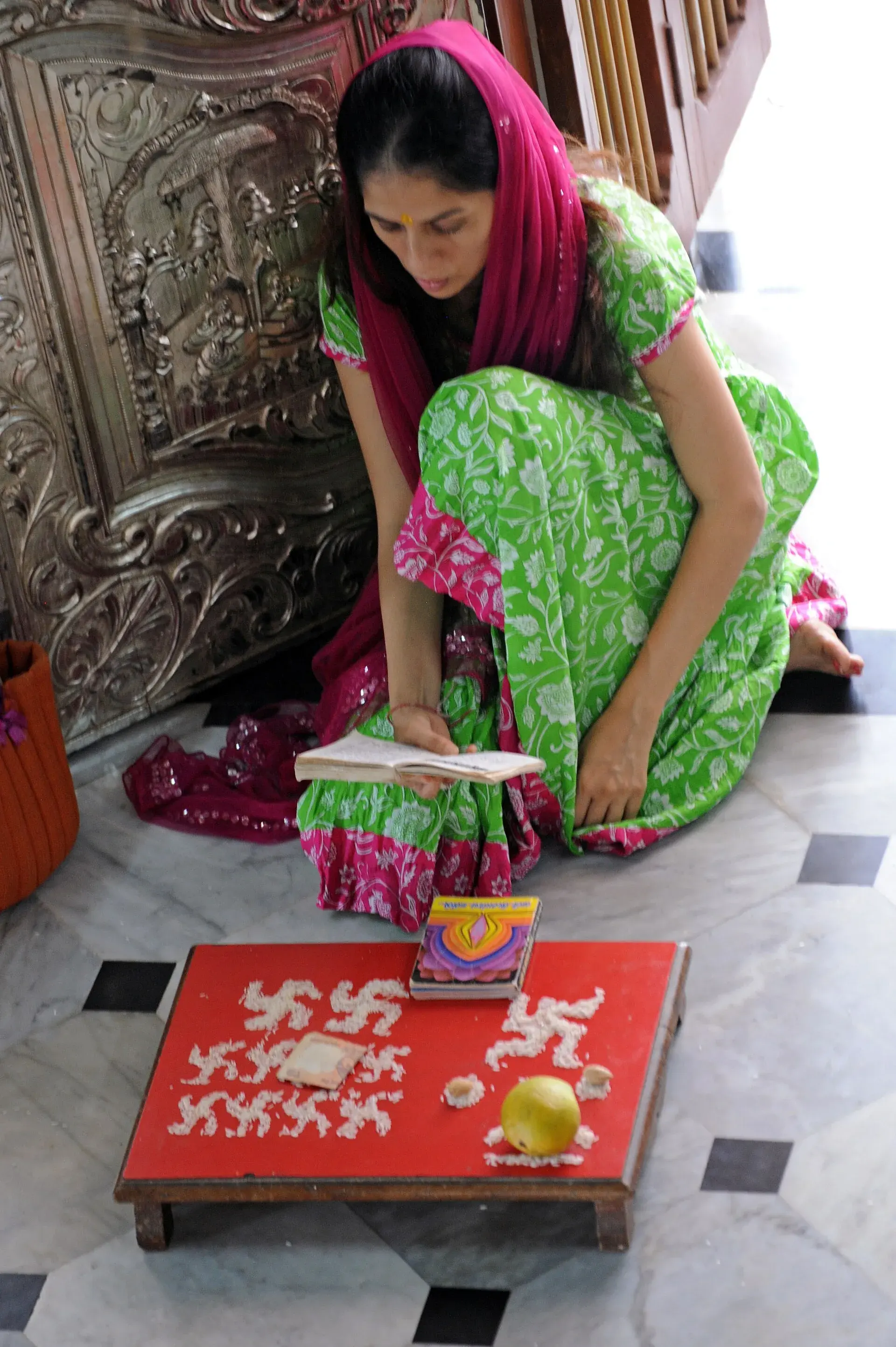 संगमरवरी फरशीवर बसलेली एक स्त्री लहान पुस्तकातून पाठ करत आहे. समोरच्या खालच्या लाल चौरंगावर अक्षतांनी स्वस्तिक व इतर चिन्हे मांडली आहेत, सोबत एक लिंबू, दोन लहान अर्घ्ये व एक छापील पुस्तिका.