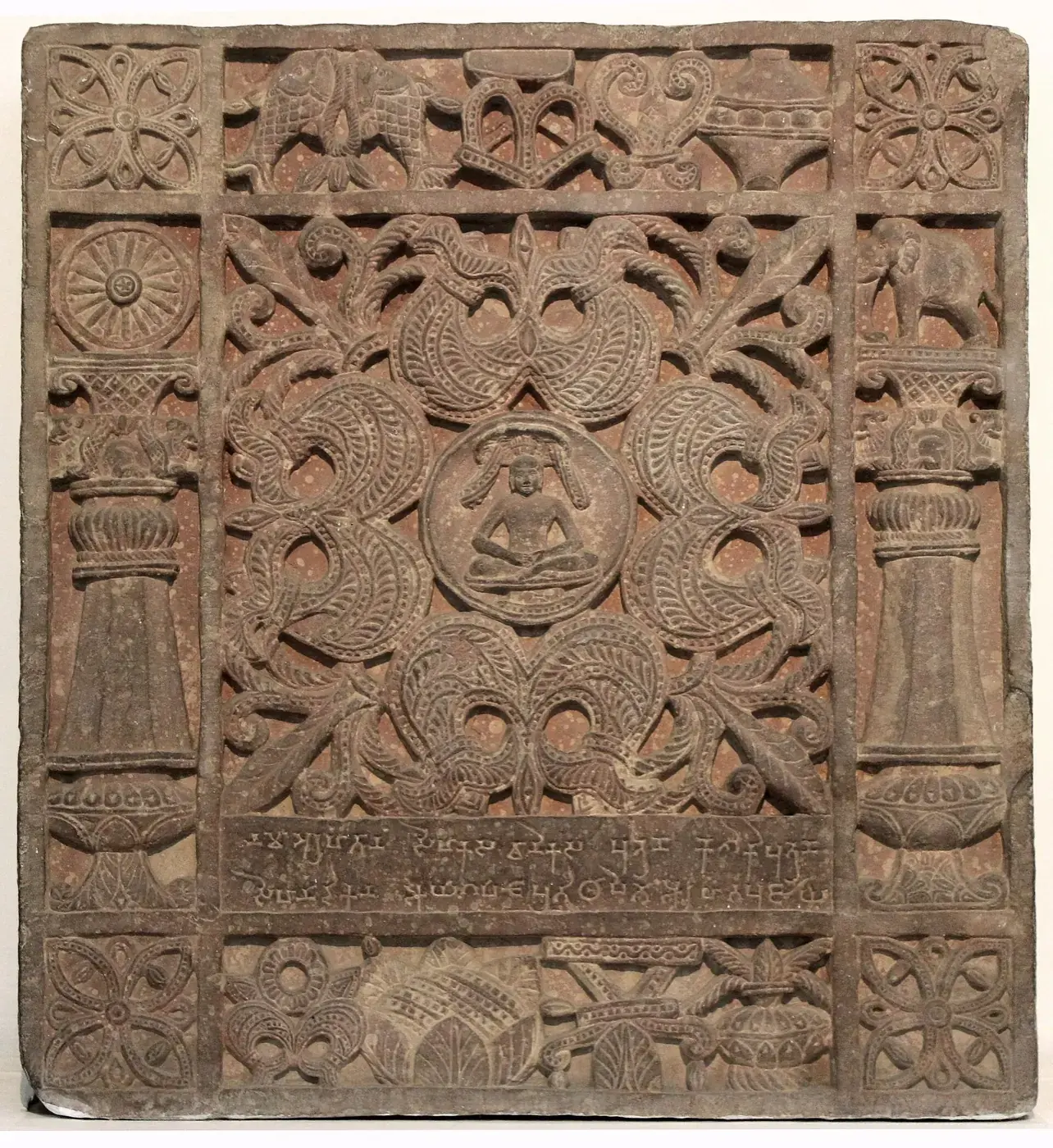 मथुरेचा कोरीव वाळूखडकाचा आयागपट, जवळपास चौरस, ज्याच्या मध्यभागी एक लहान वस्त्रहीन बसलेली जिनमूर्ती आहे, भोवती संकेंद्रित अलंकरण पट्ट्या व खालच्या कडेला शिलालेख.